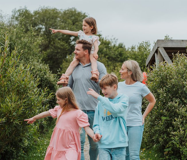 A white family of five, walk along a pathway. The youngest child, a girl, is sat on top of her father's shoulders. All five members of the family seem to be looking and pointing to something in the distance.