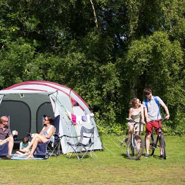 
                A pitched up family tent with a man, woman and baby sitting in the porch area of the tent. A teen boy and girl are cycling by on a hot sunny day.
                