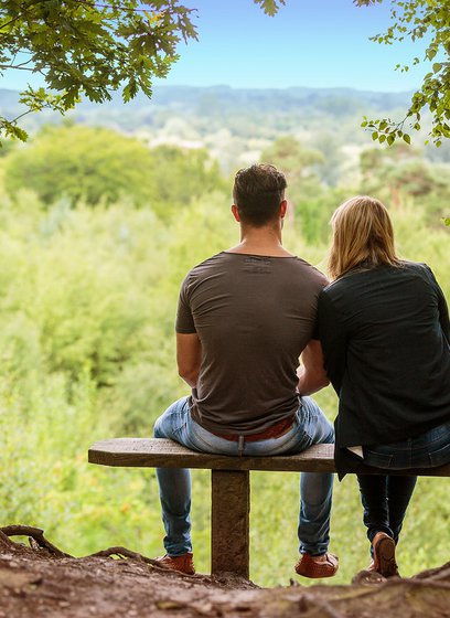 A couple sat on a bench looking out to stunning woodland views on a summers day.