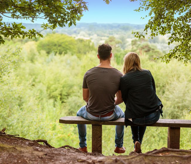 A couple sat on a bench looking out to stunning woodland views on a summers day.