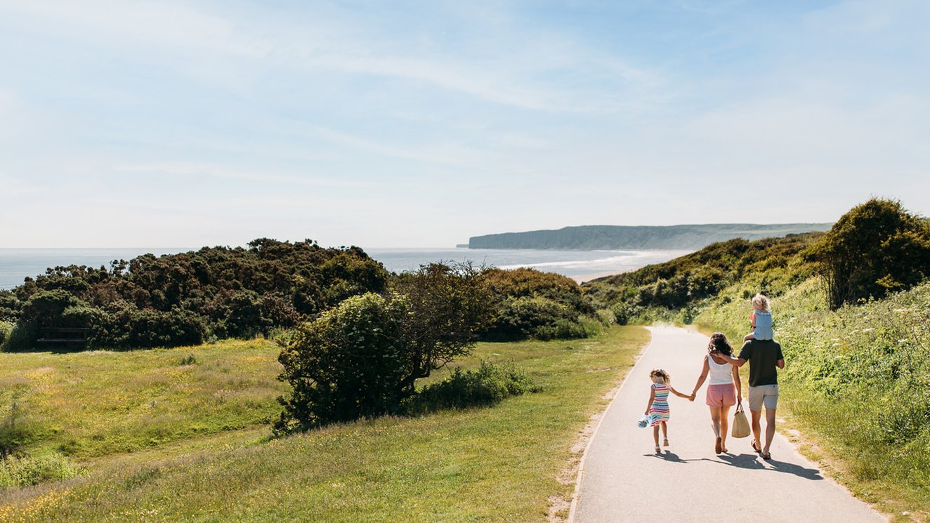 
              A young family are walking along a dry dirt path which is situated between rolling green hills on a hot summer's day.
              