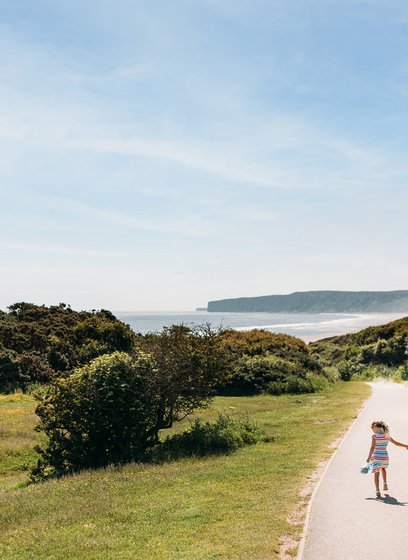 A young family are walking along a dry dirt path which is situated between rolling green hills on a hot summer's day.