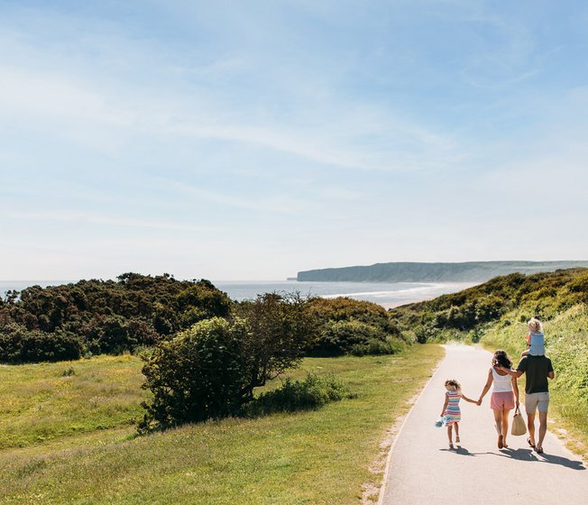 A young family are walking along a dry dirt path which is situated between rolling green hills on a hot summer's day.