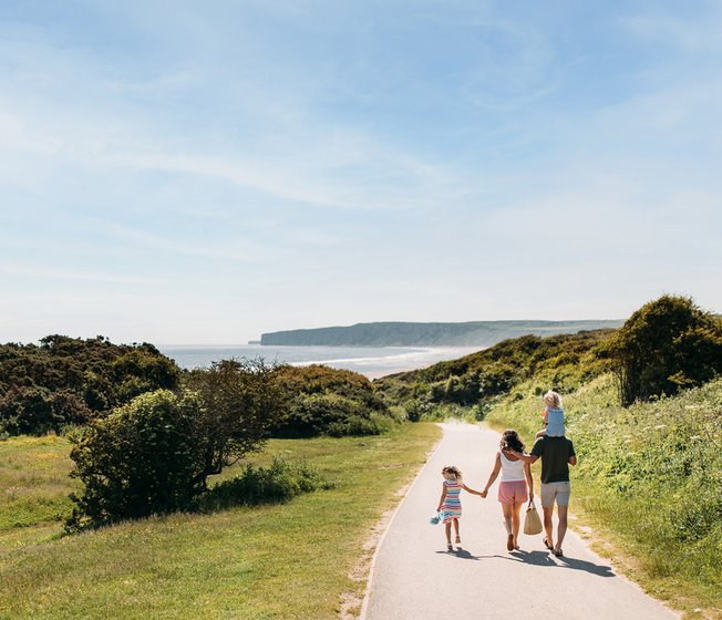 A young family are walking along a dry dirt path which is situated between rolling green hills on a hot summer's day.