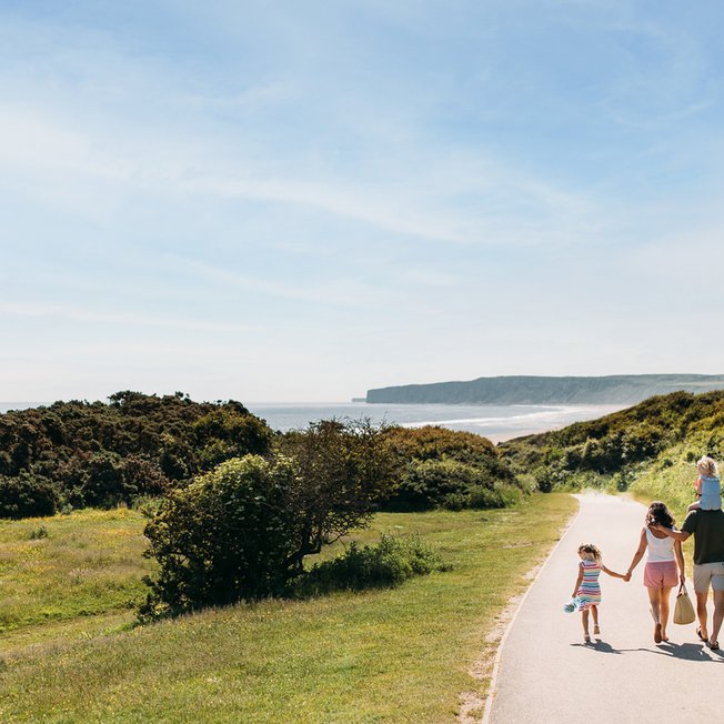 
                A young family are walking along a dry dirt path which is situated between rolling green hills on a hot summer's day.
                