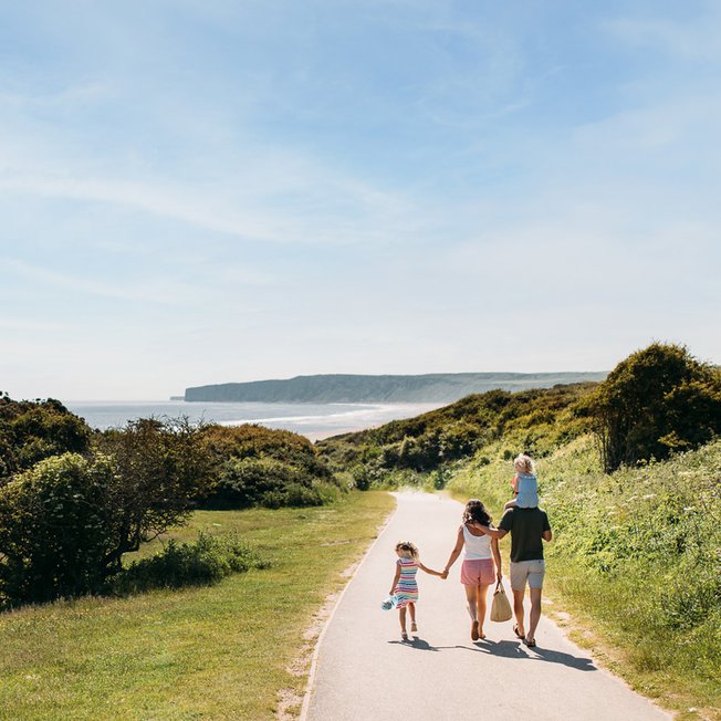 
                A young family are walking along a dry dirt path which is situated between rolling green hills on a hot summer's day.
                