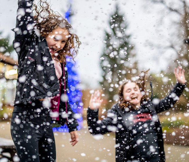 Two girls with long brown hair wearing navy blue coatsare jumping in the air excitedly and laughing as the heavy snowfall lands around them.
