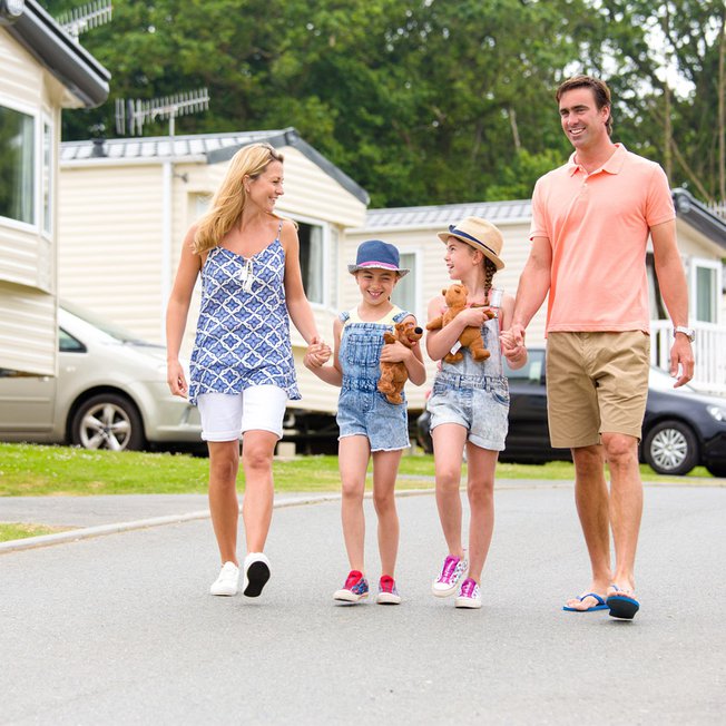 
                A family holding hands walking through one of our stunning holiday pars. There is a row of caravans in the background.
                