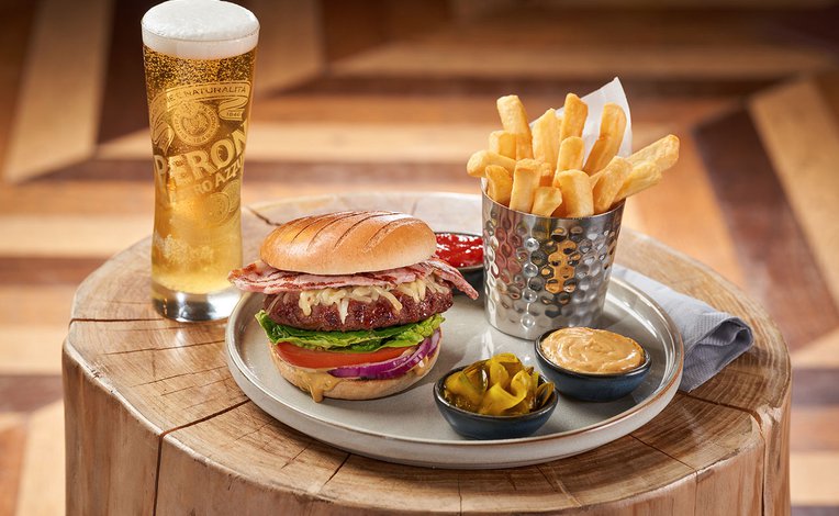 On the place is a stacked bacon cheeseburger with lettace, tomato and sliced red onion. Next to the burger is cooked chips and ketchup, pickles and burger sauce. Next to the plate on the left is a tall glass of larger with a thick, creamy head.