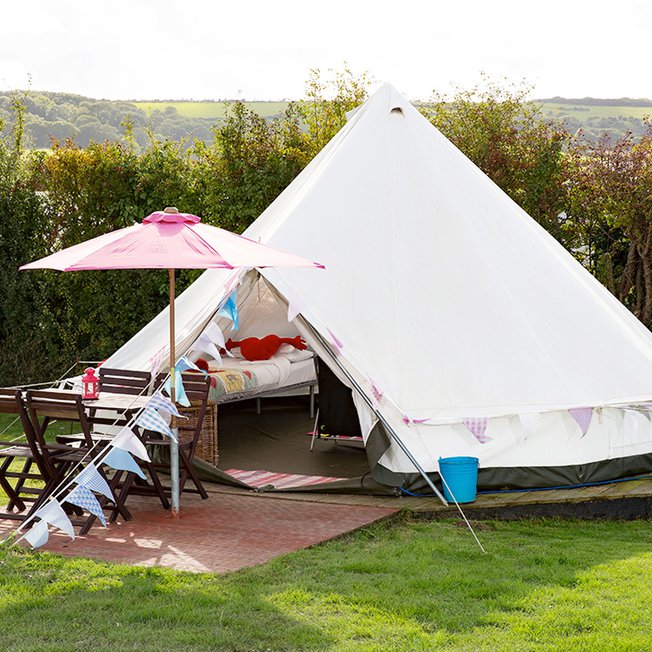
                An off white canvas bell tent pitched up outside on the grass on a sunny day. The entrance to the bell tent is open and there is a pink parasol resting against the entrance.
                