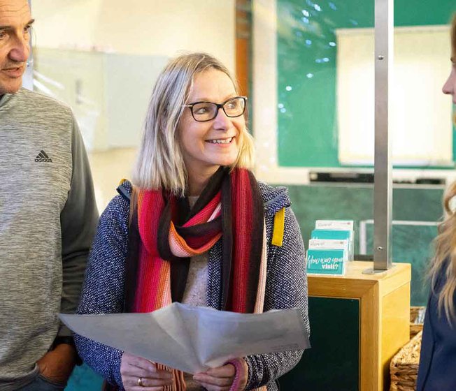 A photo of a mature man and woman mid sentence. The man has long grey hair and is wearing a grey pullover jumper. The woman also has long grey hair and is wearing a scarf, glasses and a dark blue hoodie. She is holding a map.