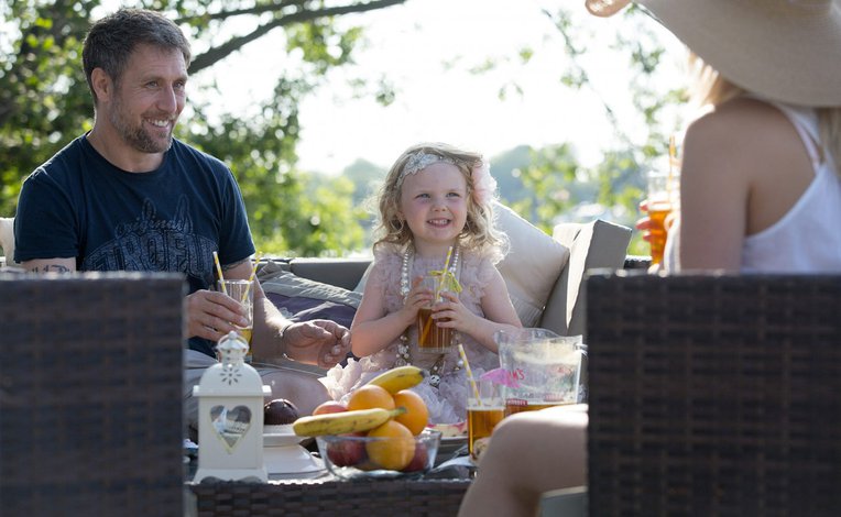 A family with a young child sitting outside enjoying a drink. On the table is a bowl of oranges and bananas.