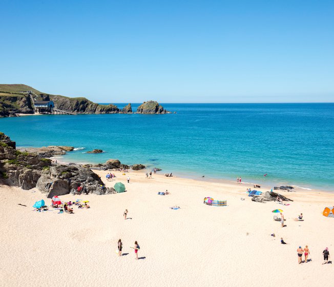 An aerial view of Padstow beach. Holiday goers are on the beach and in the sea. A bright blue sky is above the sea.