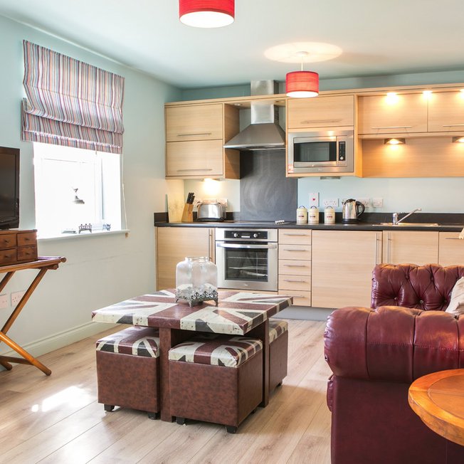 
                An open-plan view of one of our apartments. A wooden modern kitchen spreads across the back wall, with a red leather sofa on the right side, a compact four-seater table in the middle with a TV opposite the sofa.
                