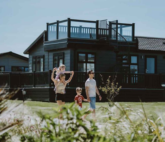 A young family taking a stroll past our stunning holiday homes with roof terraces. One of the two children is on their mum's shoulders.