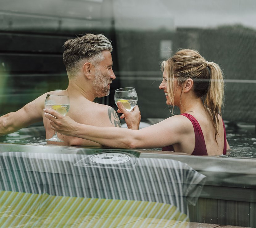 A middle-aged couple are cuddled with drinks in hand, whilst sitting in a hot tub.